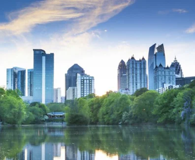 Atlanta skyline reflected in a calm lake surrounded by trees under a blue sky