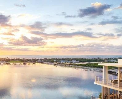 Rendering of a curved building balcony with furnished terraces overlooking a wide waterway at sunset, with a shoreline of trees and estates in the distance and the ocean beyond.